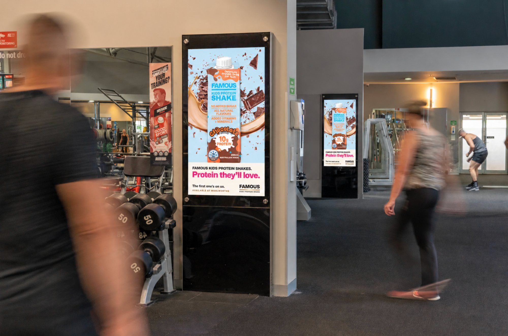 Famous kids protein shake on digital screens across a busy gym floor, members walking past